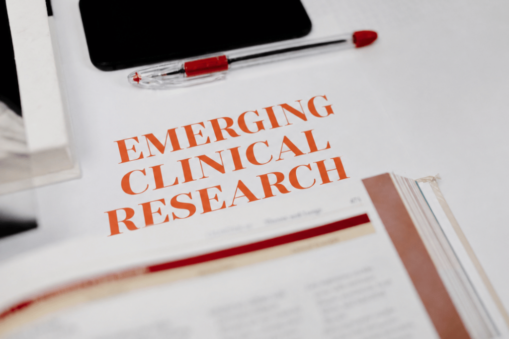 Close-up of a clinical research document titled "Emerging Clinical Research" on a desk with a pen, smartphone, and reference materials, representing the growing body of scientific evidence supporting IHHT.