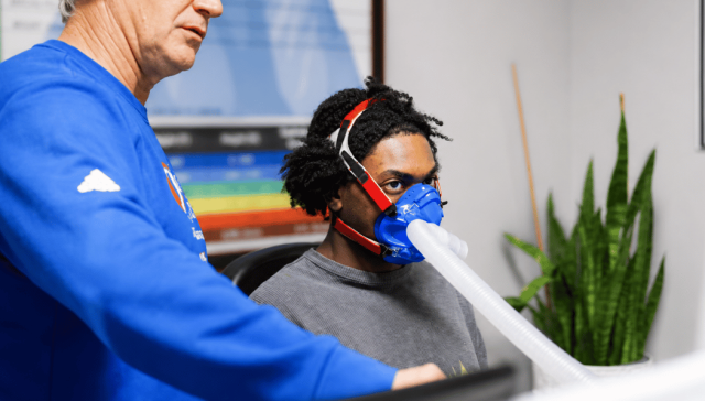 Coach guiding a client through an IHHT session while the client pedals gently on a recumbent bike wearing an oxygen training mask.