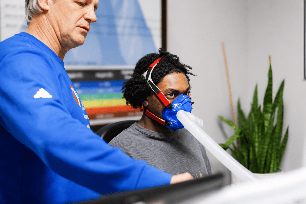 Coach guiding a client through an IHHT session while the client pedals gently on a recumbent bike wearing an oxygen training mask.