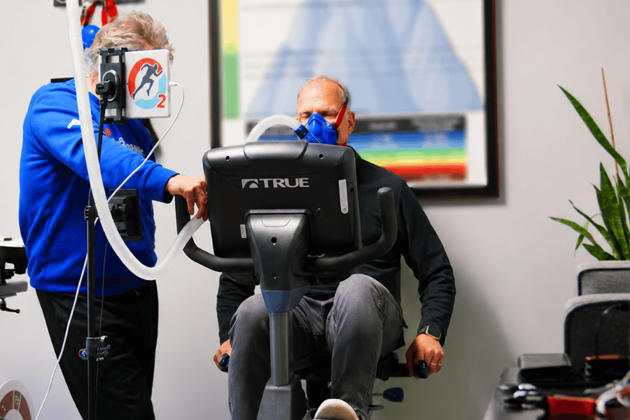 Coach supervising a client wearing an IHHT breathing mask while pedaling a stationary bike during a guided training session.